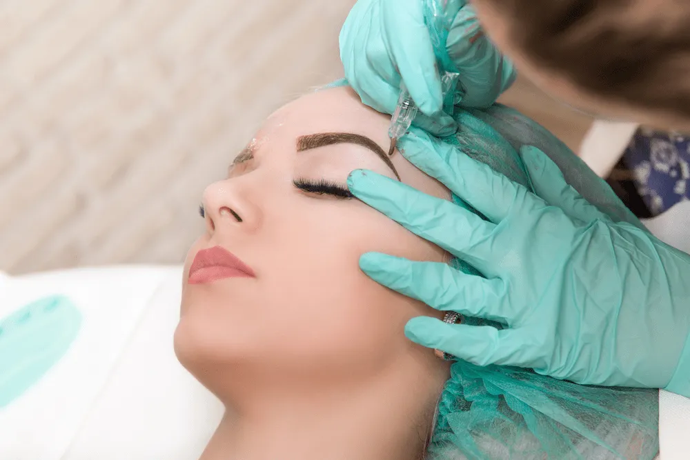 Woman laying down on a table while someone else does microblading work on her eyebrows.