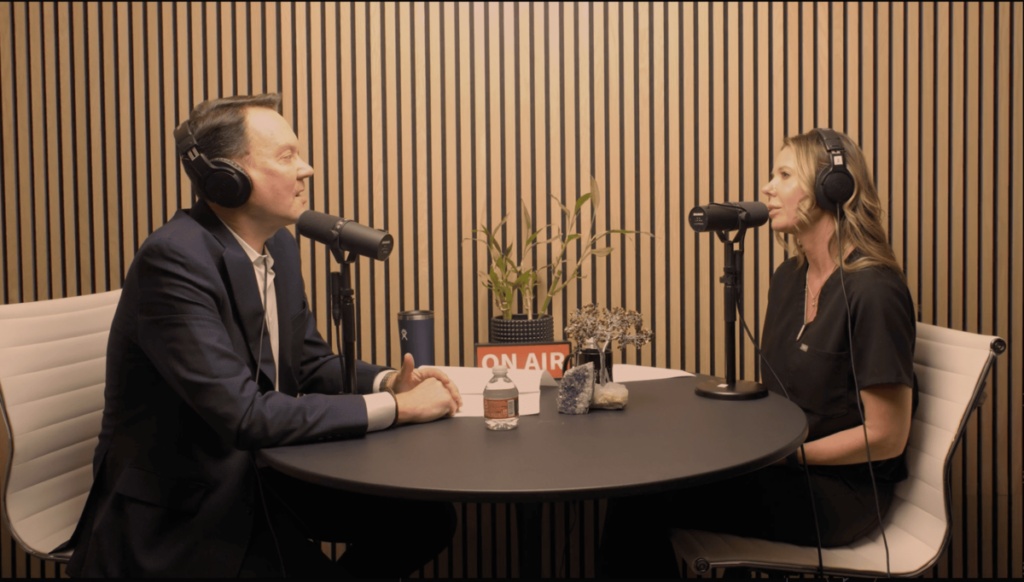 Two people wearing headphones at a podcast table with microphones and a sign that reads "ON AIR."