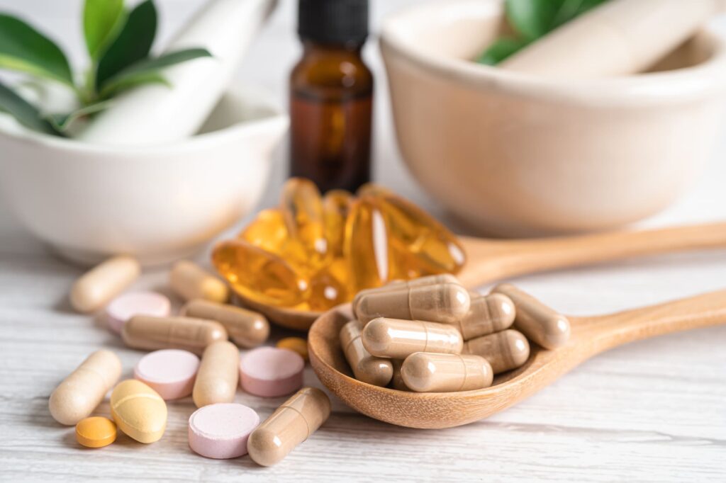 Assorted dietary supplements, including capsules, tablets, and softgels, on a wooden surface with a ceramic mortar and brown bottle in the background.