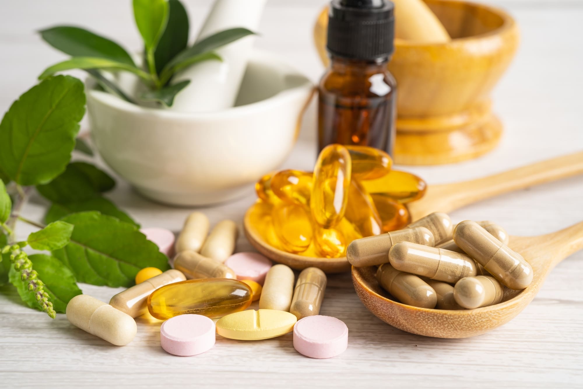 Assorted capsules, tablets, and softgels arranged with green leaves and a mortar and pestle.