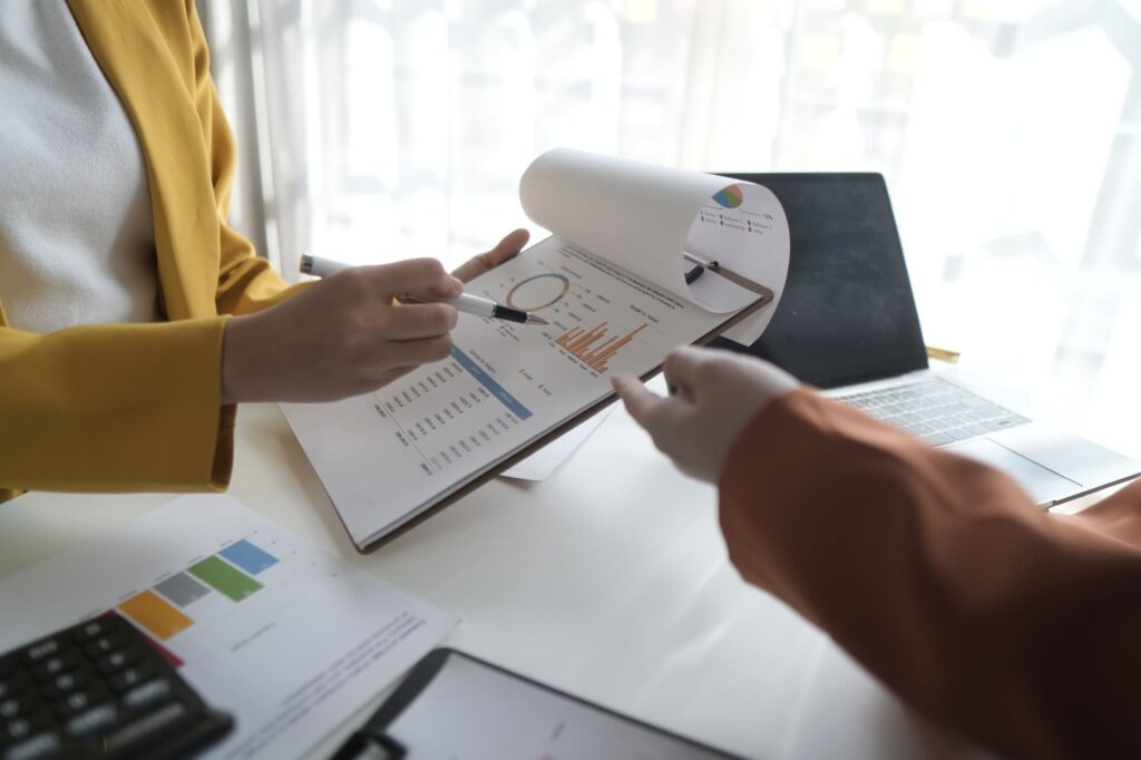 Two people discuss charts on a document at a well-lit desk with a laptop and calculator.