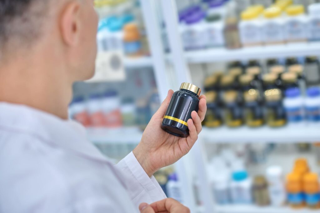 Person in a white coat examining a dark bottle with a gold cap in front of shelves filled with bottles.