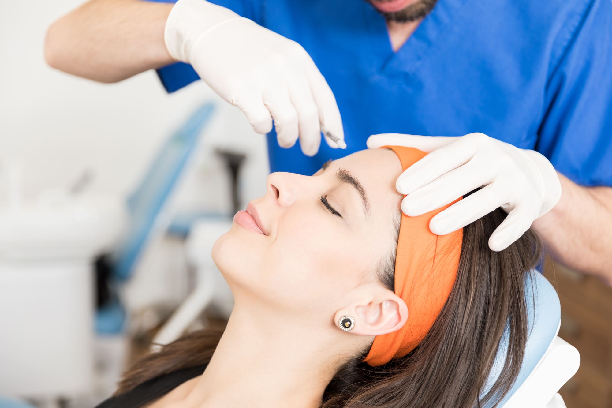 A person with an orange headband and closed eyes receives a facial treatment from someone in blue scrubs wearing gloves. The atmosphere is calm and professional.