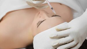 A person receiving a cosmetic injection in the forehead while lying down, wearing a headband. Gloved hands hold the syringe. Calm, clinical atmosphere.